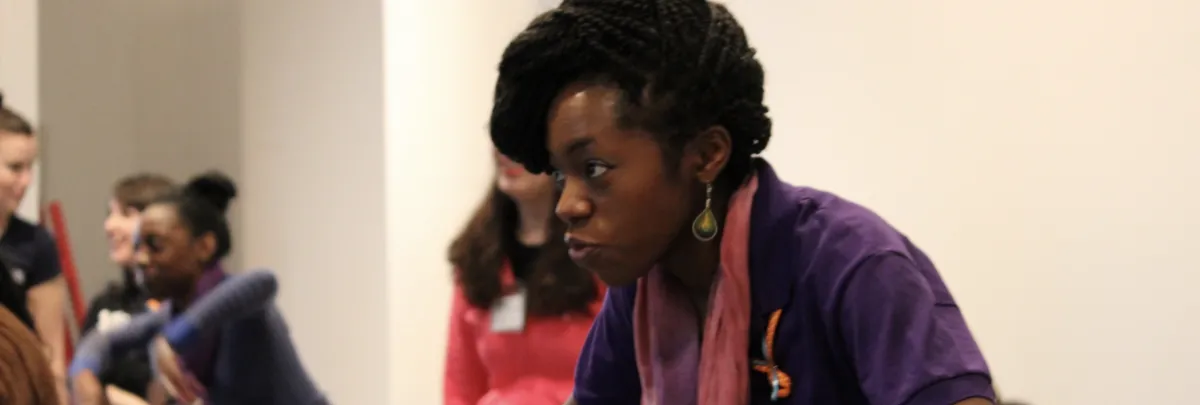 A woman in a purple shirt and pink scarf leans forward, appearing focused, with several people blurred in the background.