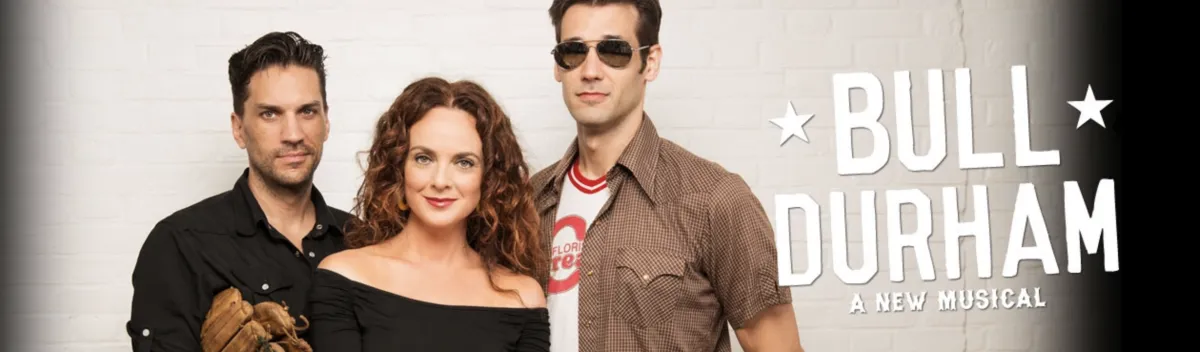Three people pose in front of a white brick wall with the text "Bull Durham: A New Musical" displayed beside them.