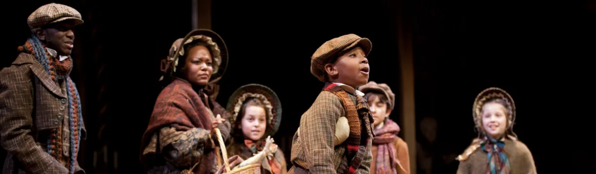 A group of people in period costumes stand on stage, with one boy at the center appearing to speak or sing while others watch him.