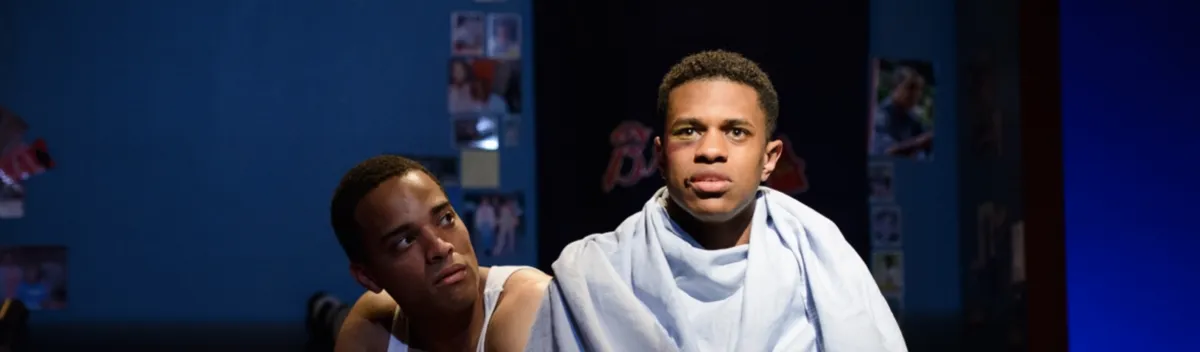 Two young men sit indoors; one is covered with a light blue sheet and looking forward, while the other looks at him. The background is dimly lit with photos on the wall.