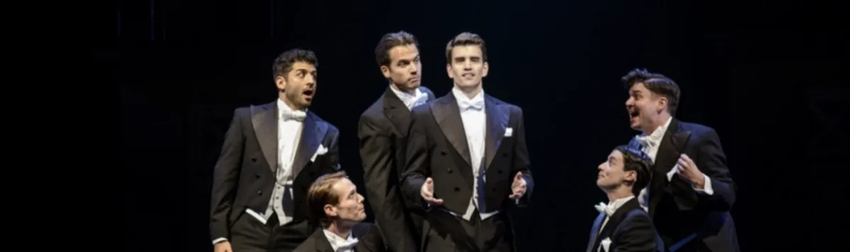 Six men in tuxedos perform on stage, singing or speaking with expressive gestures against a dark background.