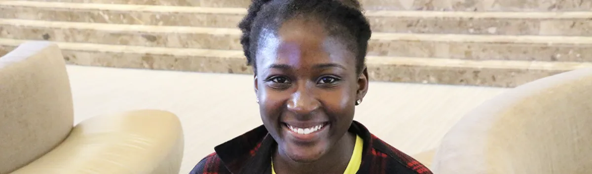 A person with dark hair pulled back is smiling while sitting indoors on a light-colored chair, with marble steps in the background.