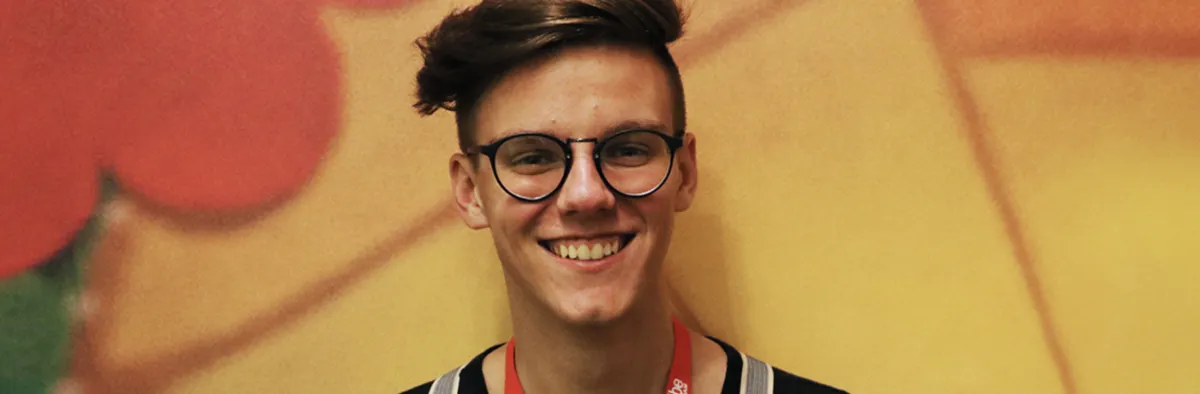 A young person with short brown hair, glasses, and a red lanyard smiles in front of a colorful, abstract background.