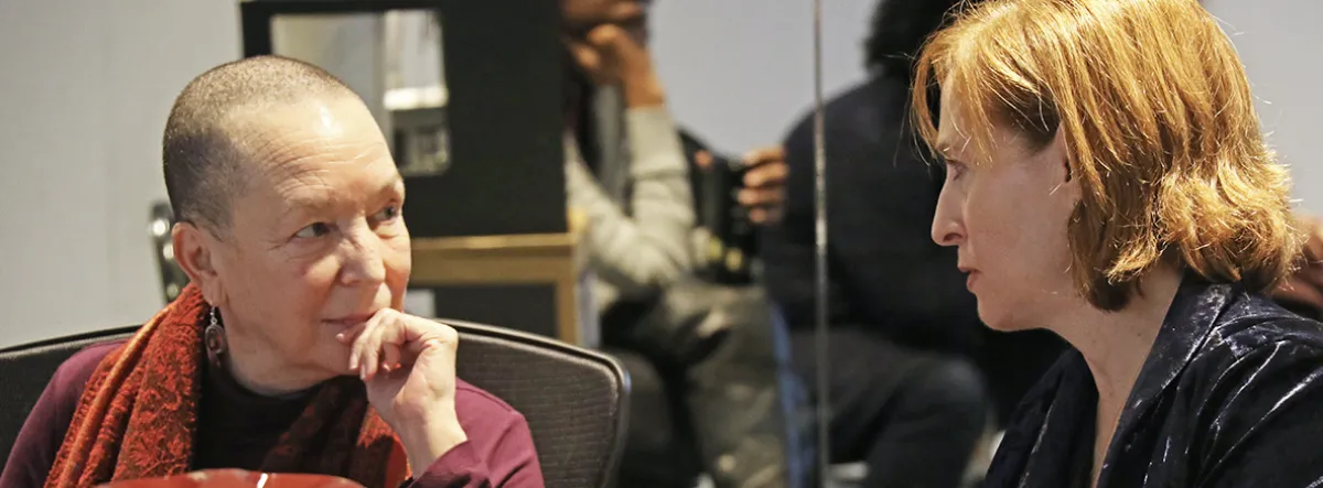 Two women sit facing each other and have a conversation in an indoor setting. One has very short hair and a red scarf; the other has light brown hair and is wearing a dark jacket.