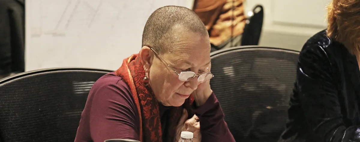 A person with short hair and glasses reads or looks down at papers while seated at a table, wearing a maroon shirt and patterned scarf.