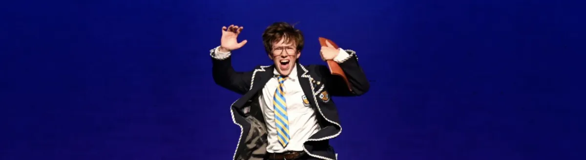 A person in a school uniform and striped tie stands on stage with arms raised, holding a book, against a plain blue background.
