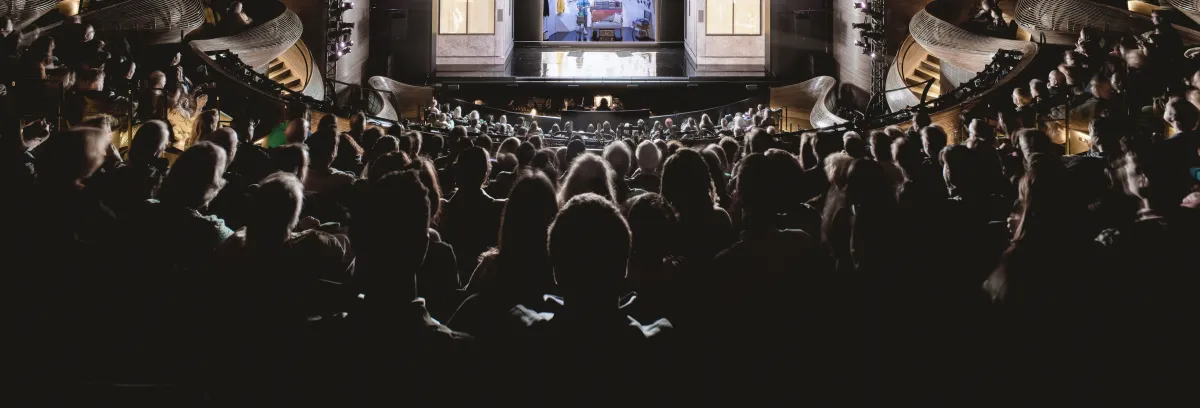 A large audience sits in a dark theater facing a brightly lit stage with performers and a screen displaying an image.