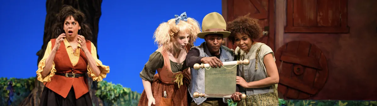 Four actors in colorful costumes perform on stage, with three reading from a scroll and one looking surprised; a painted backdrop suggests a whimsical setting.