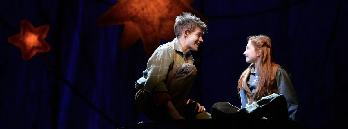 Two young actors sit on stage under star-shaped lights, facing each other and smiling, with a dark blue background behind them.