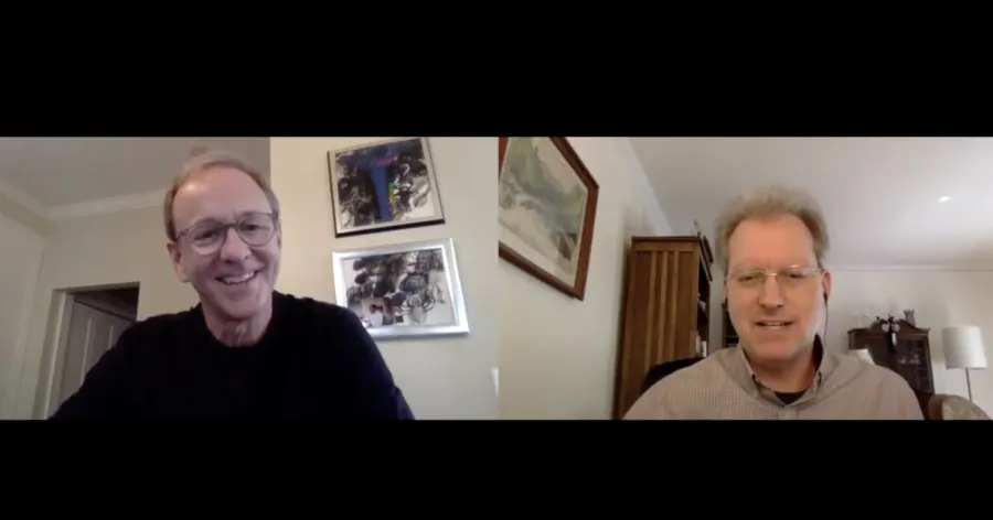 Two men have a video call, each sitting in separate rooms with art and furniture visible in the background.