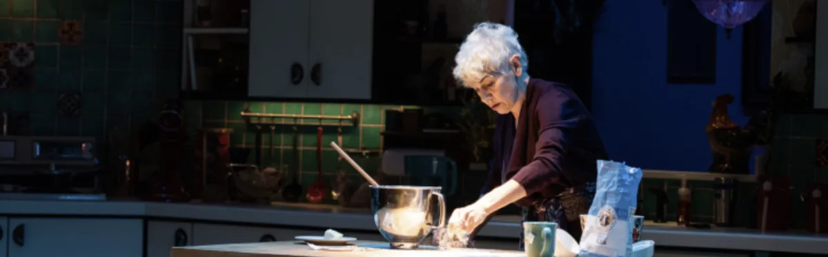 An older woman stands at a kitchen counter preparing food, with a mixing bowl, a mug, and ingredients on the counter under focused lighting.