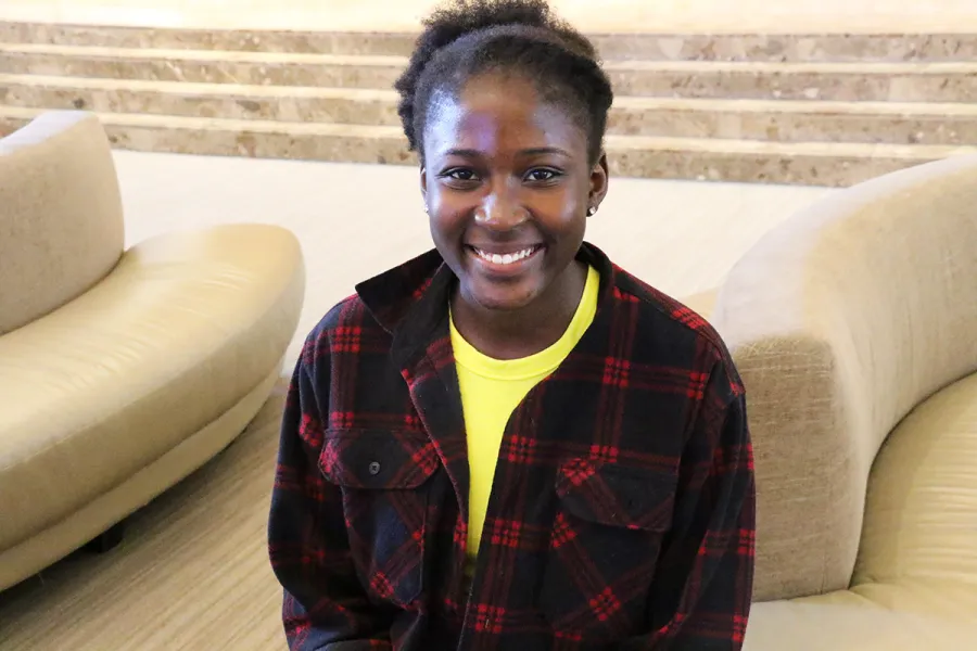 A person wearing a black and red plaid shirt over a yellow top sits indoors on a beige chair, smiling at the camera.
