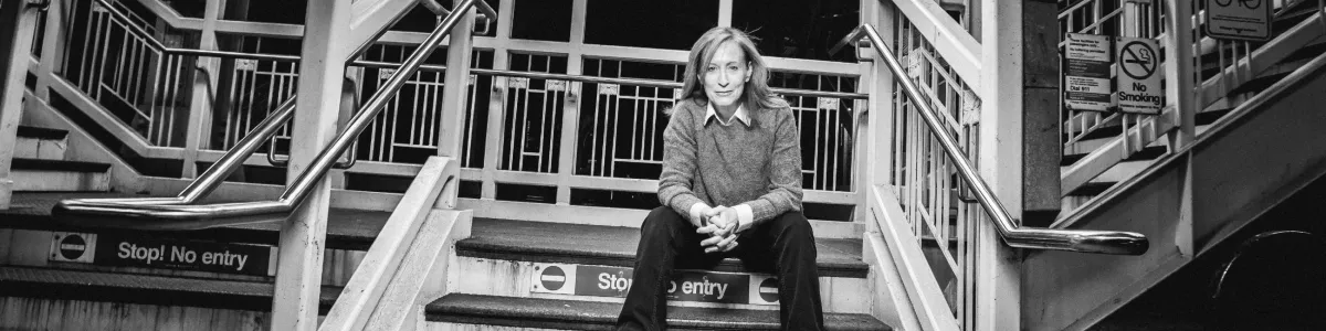 A person sits on stairs in front of "Stop! No entry" signs, surrounded by metal railings and multiple warning notices.