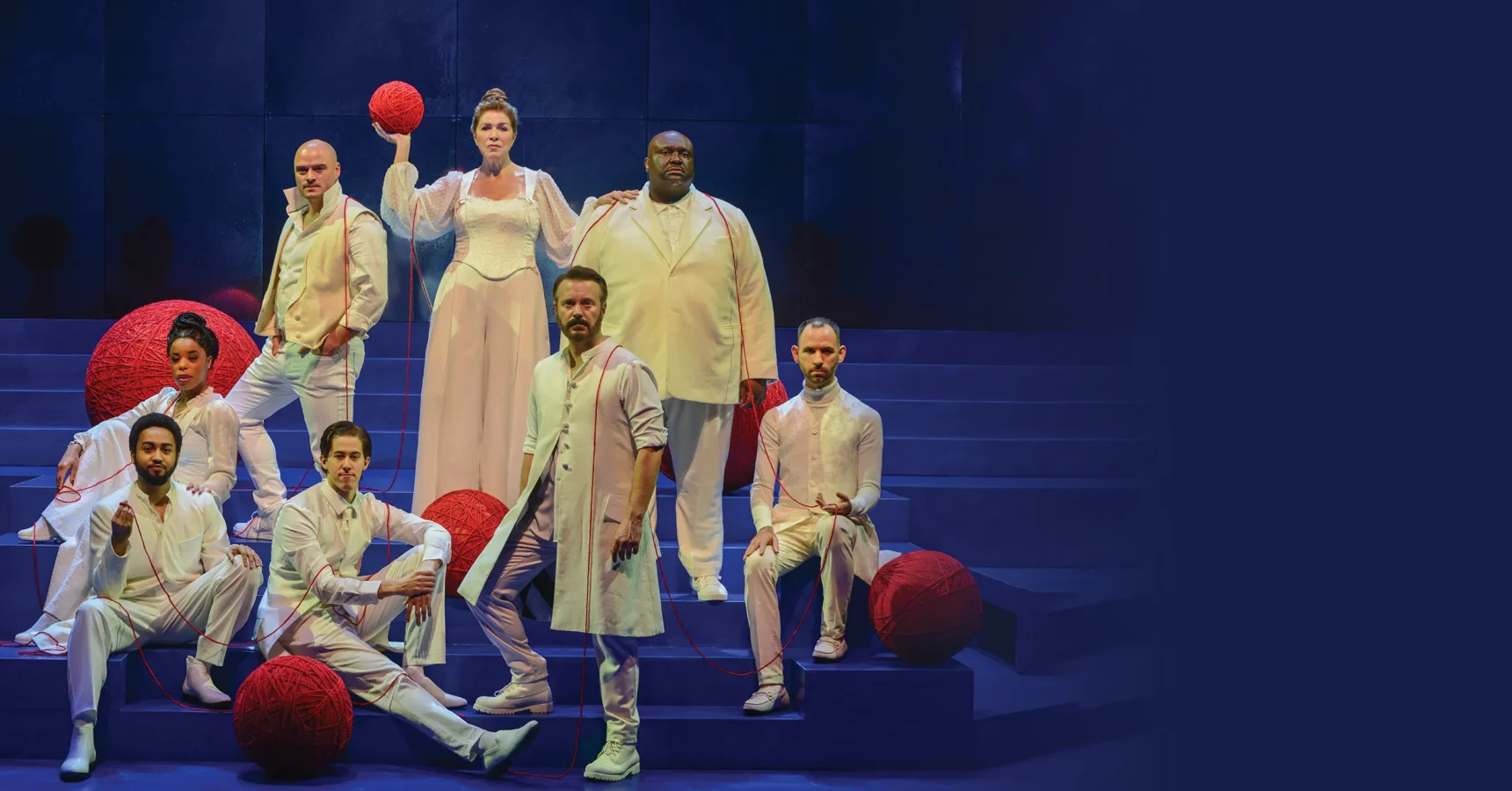 Seven people in white attire pose on blue steps, surrounded by large red yarn balls, against a dark blue background.