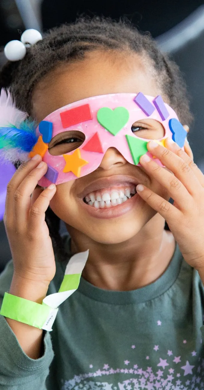 Child smiling and holding a decorated paper mask with colorful shapes and feathers, wearing a green shirt and a wristband.