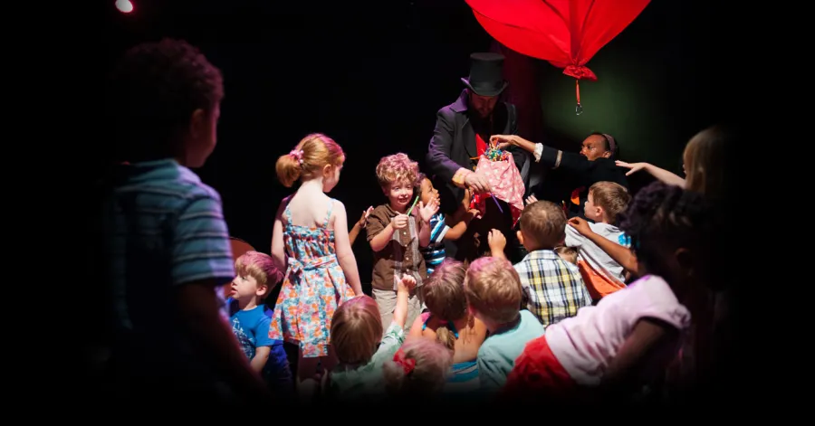 A group of children surround a magician wearing a top hat who is performing a trick on stage under a large red balloon.