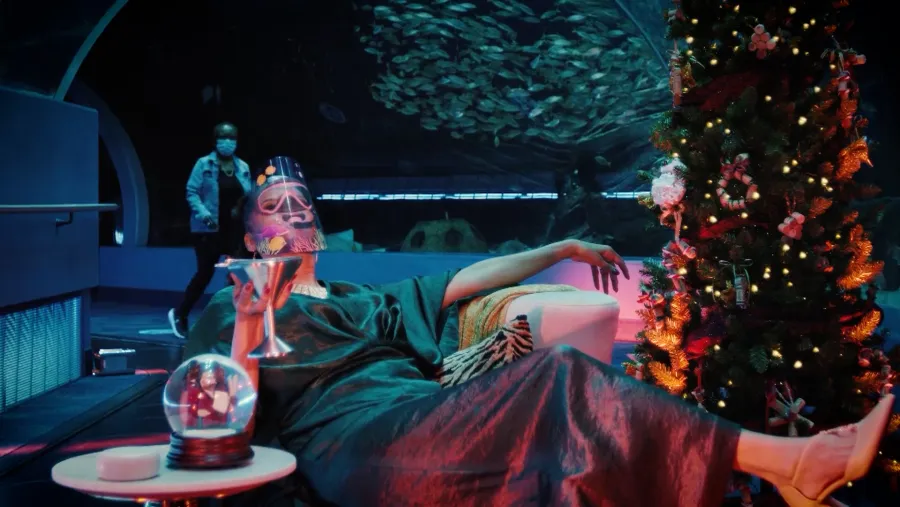 A person in a robe and mask lounges with a cocktail near a decorated Christmas tree in an aquarium, while another person walks in the background.