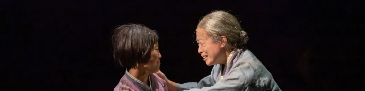 Two people in traditional clothing face each other closely, one smiling warmly at the other, against a dark background.