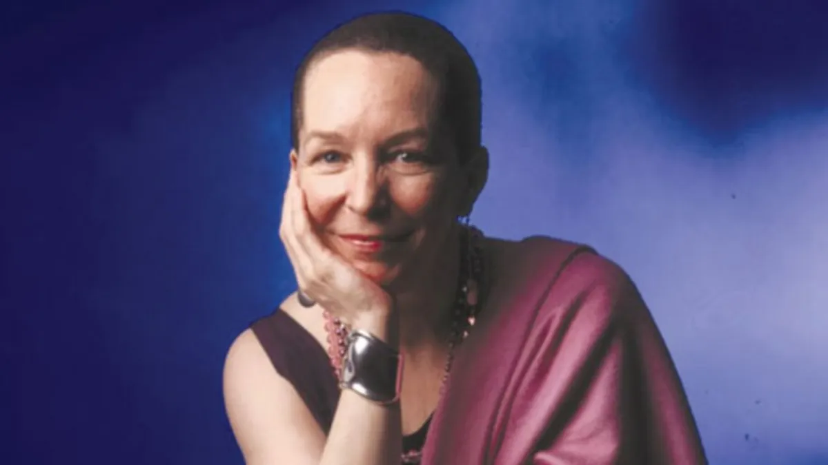 A woman with short hair and a slight smile rests her head on her hand, wearing a sleeveless top, a shawl, and jewelry, against a blue background.