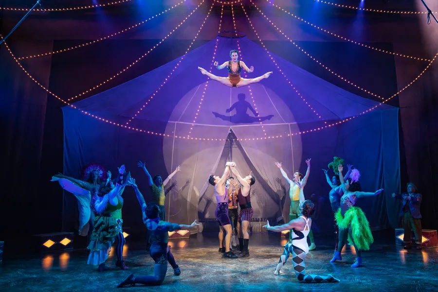 A group of circus performers are on stage; three acrobats lift a person while another acrobat performs a split midair above them under stage lights.