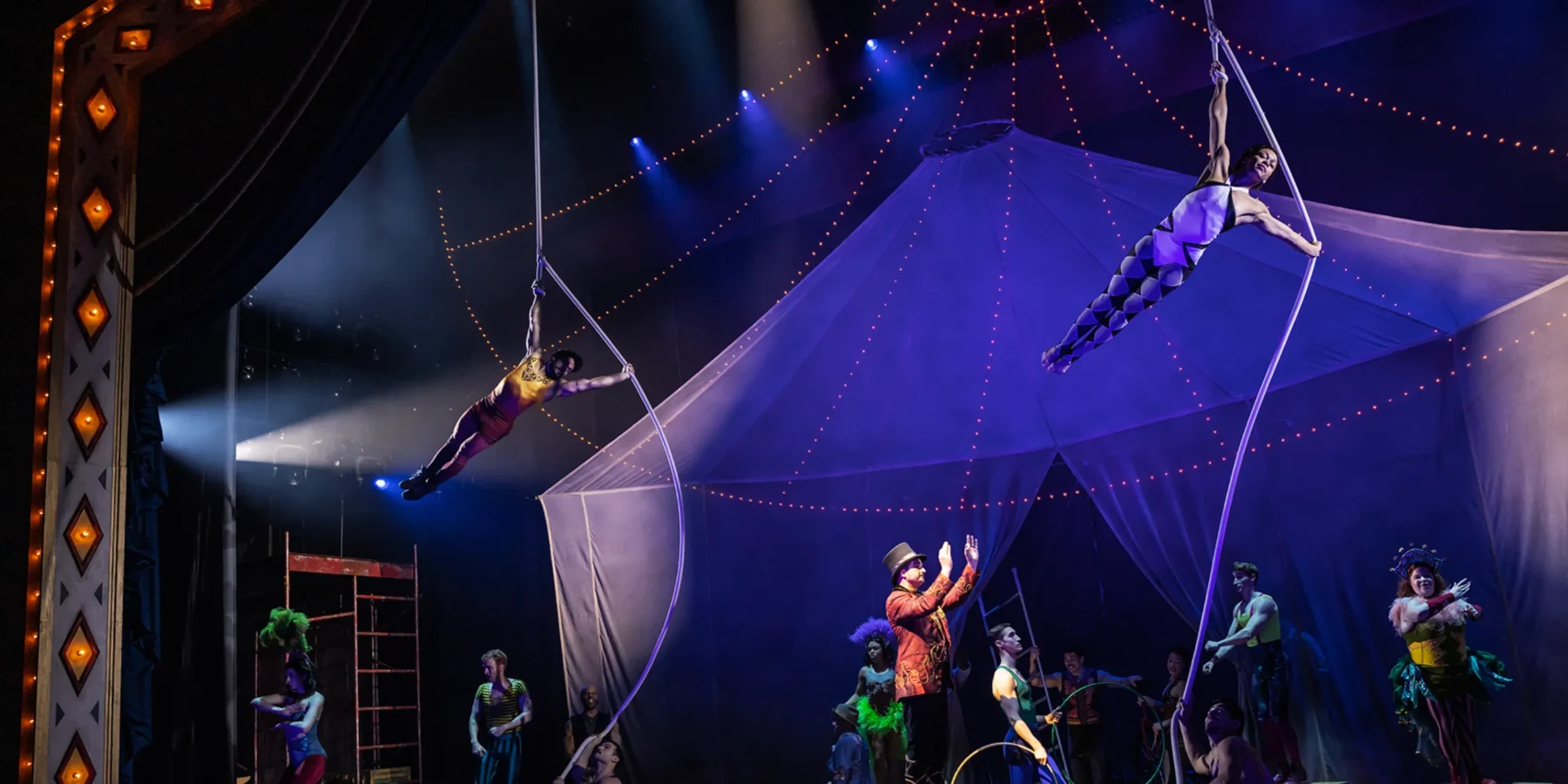 Circus performers on stage with acrobats suspended on ropes above, a ringmaster below, and other costumed artists in front of a large, tent-like backdrop under stage lights.
