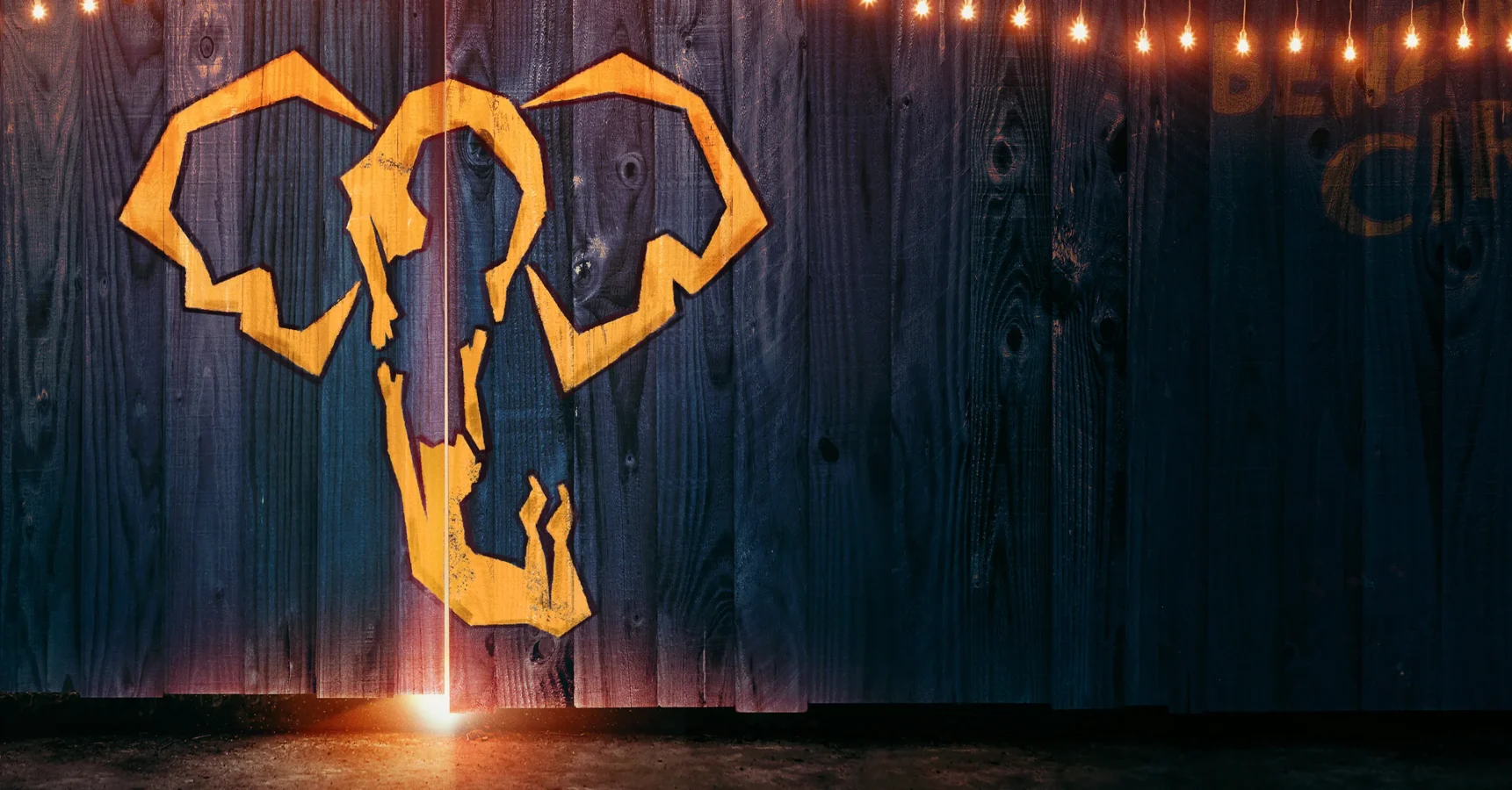 A stylized yellow elephant head is painted on blue wooden boards, illuminated by a row of small lights along the top edge.