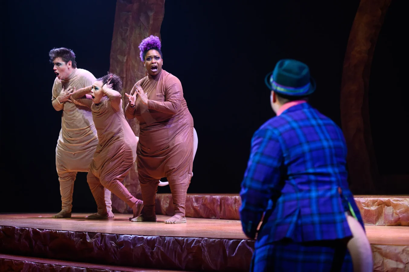 Three performers in tan costumes strike animated poses on stage while facing a person in a blue plaid suit and green hat. The background is dark with minimal props.