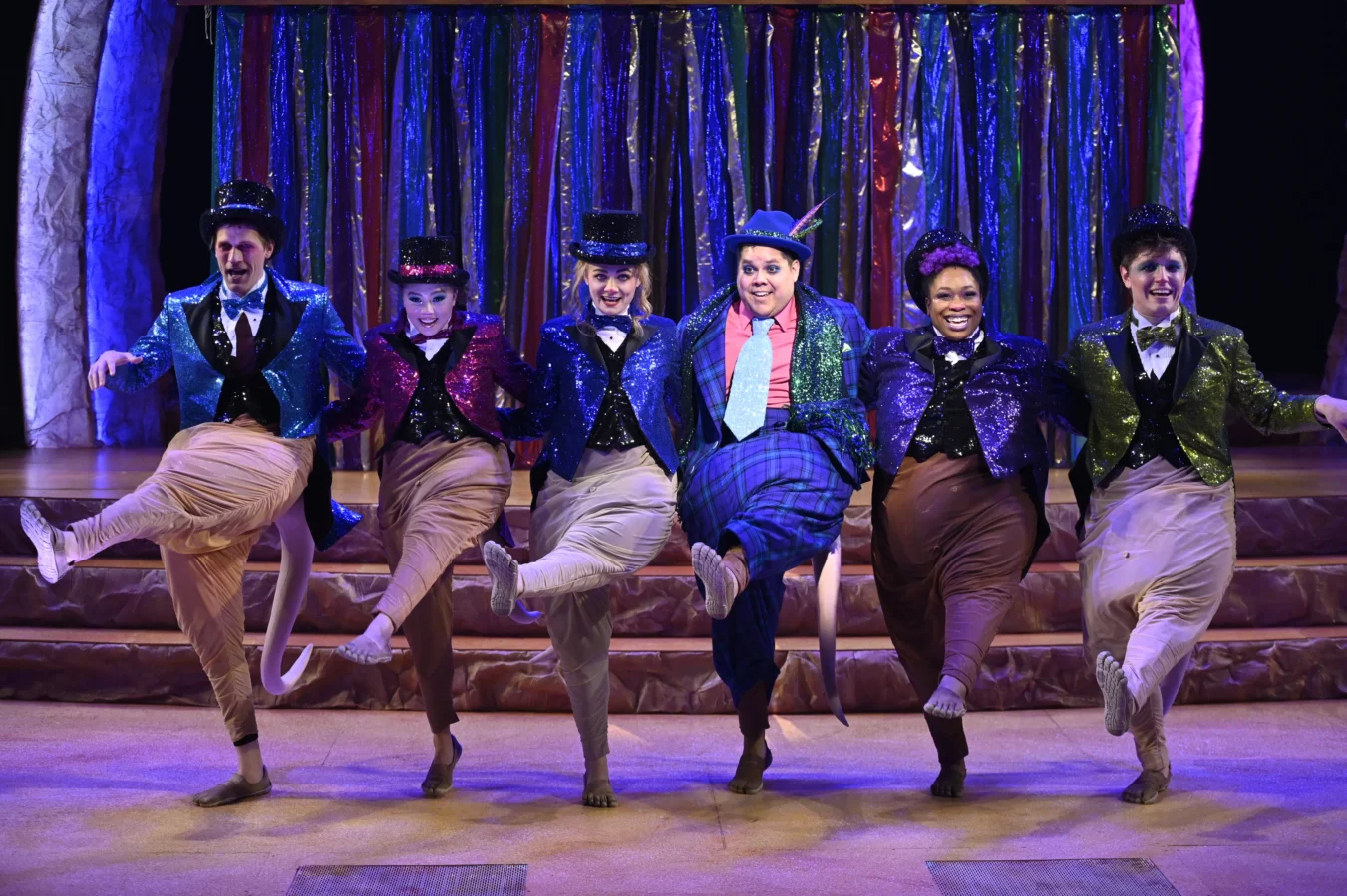 Six performers in colorful, sequined costumes and top hats dance in a line on stage, each lifting one leg, with a curtain of metallic streamers in the background.