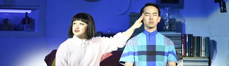 A woman with a bob haircut touches the head of a man with closed eyes, both sitting indoors under blue lighting, with shelves and books in the background.
