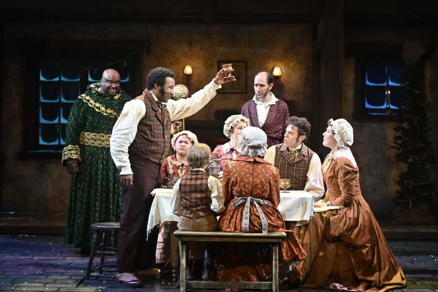 A group of actors in period costumes gather around a table on stage, with one man standing and raising a glass while others watch him.