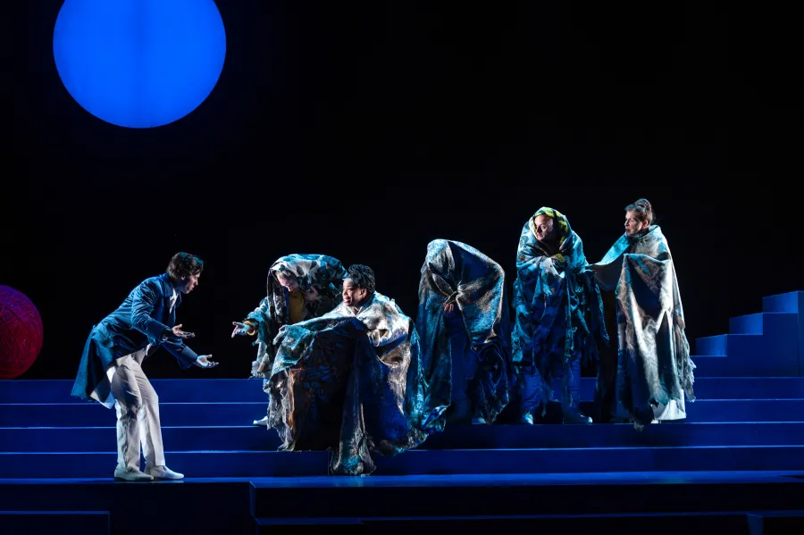 A man in blue and white gestures toward five people draped in metallic robes, standing on blue steps under a large blue circle light.