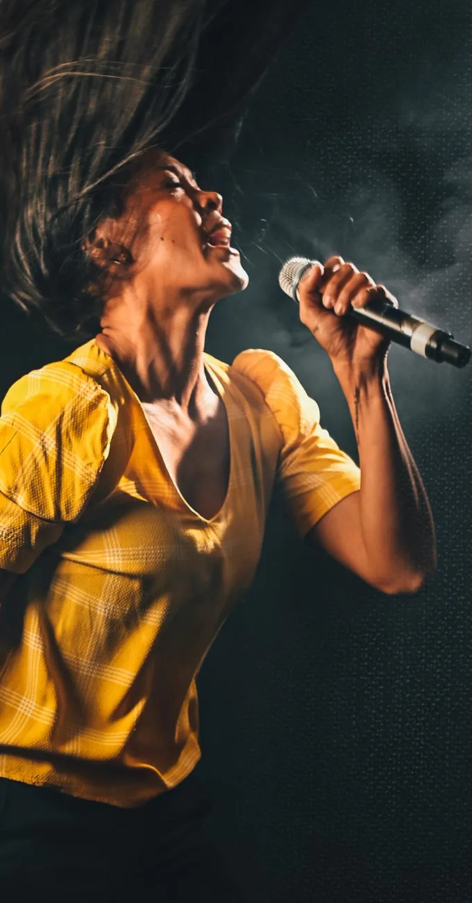 Person wearing a yellow shirt passionately singing into a microphone on stage, with hair flying and dramatic stage lighting.