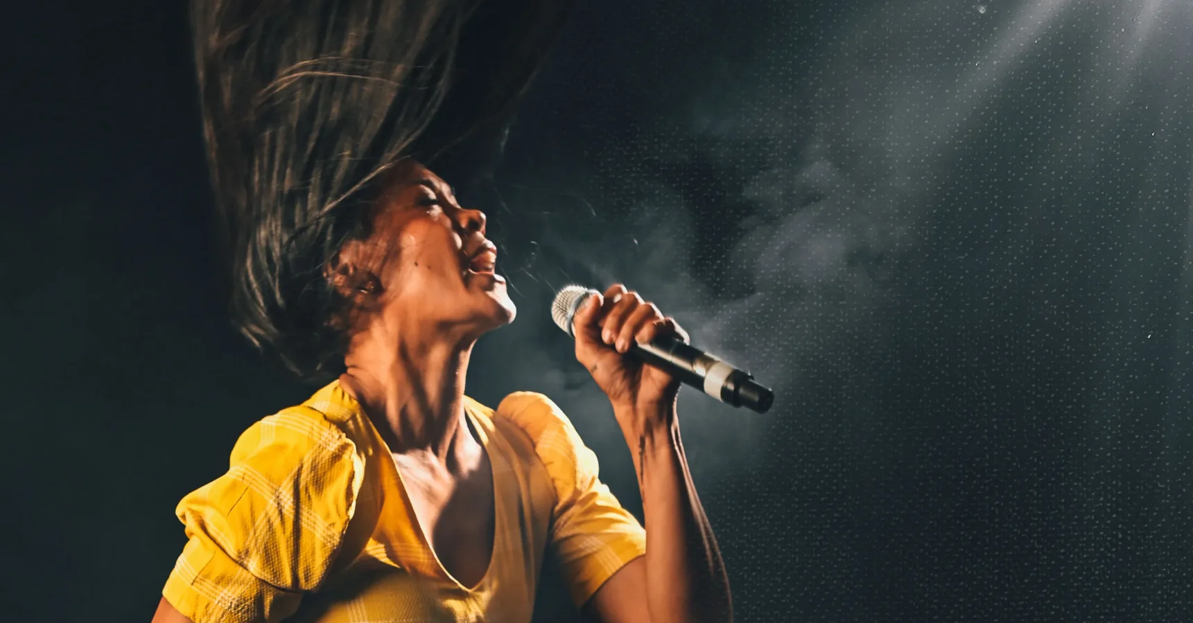 A person in a yellow shirt sings passionately into a microphone, with hair flying and dramatic stage lighting in the background.