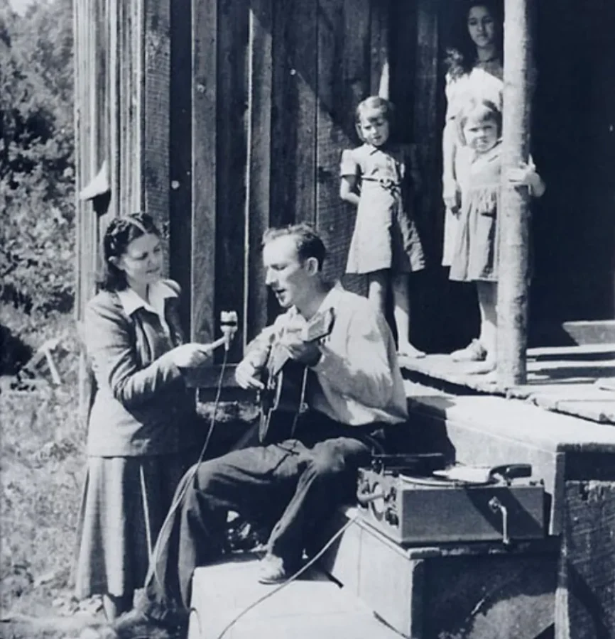 A woman holds a microphone to a seated man playing guitar on a porch, while three children stand nearby.