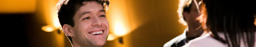 A man with curly hair smiles while looking at someone in the foreground; warm yellow lighting and blurred background.