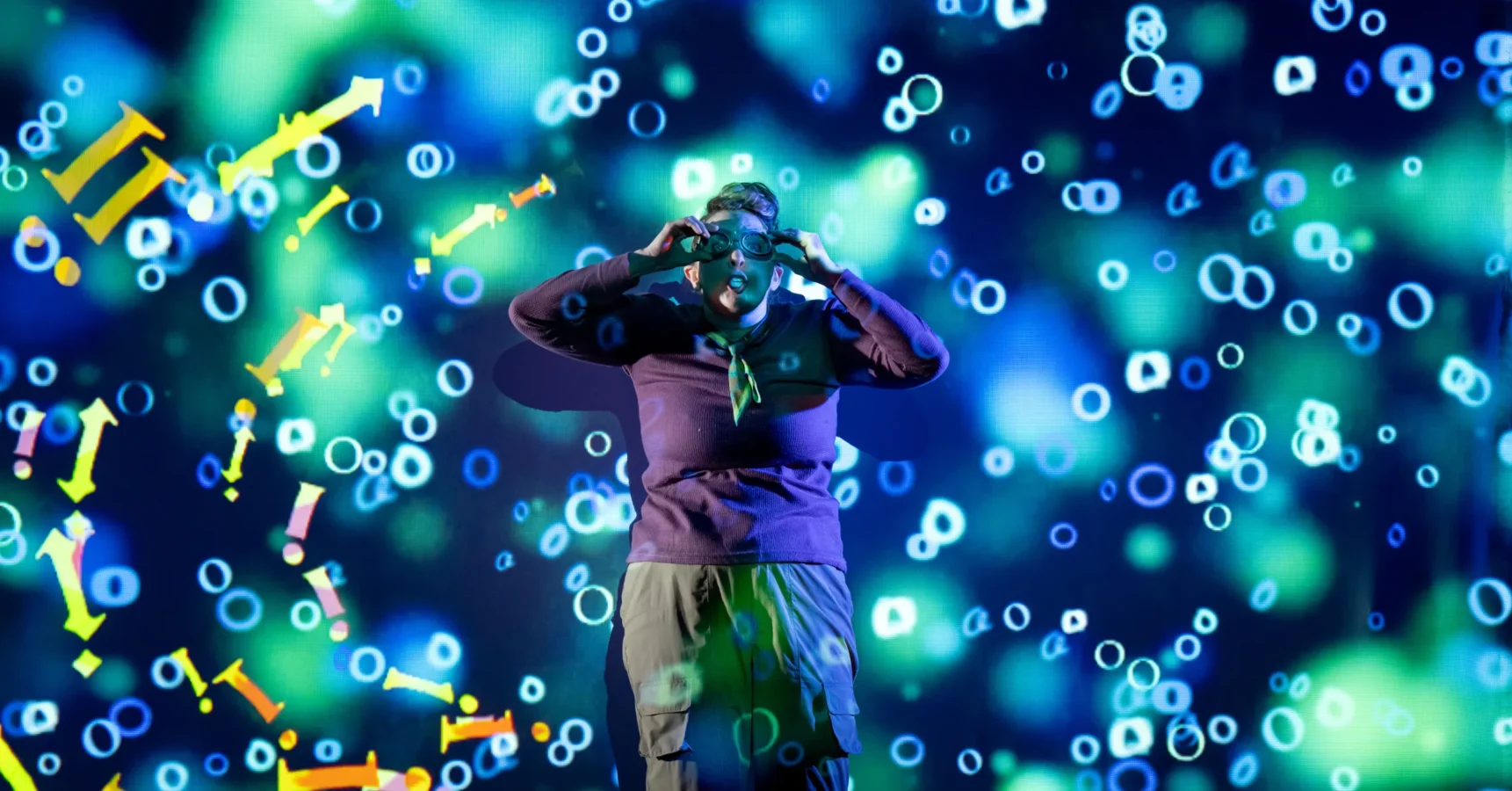 A person wearing virtual reality goggles stands in front of a digital backdrop with blue, green, and yellow circular and arrow-shaped graphics.