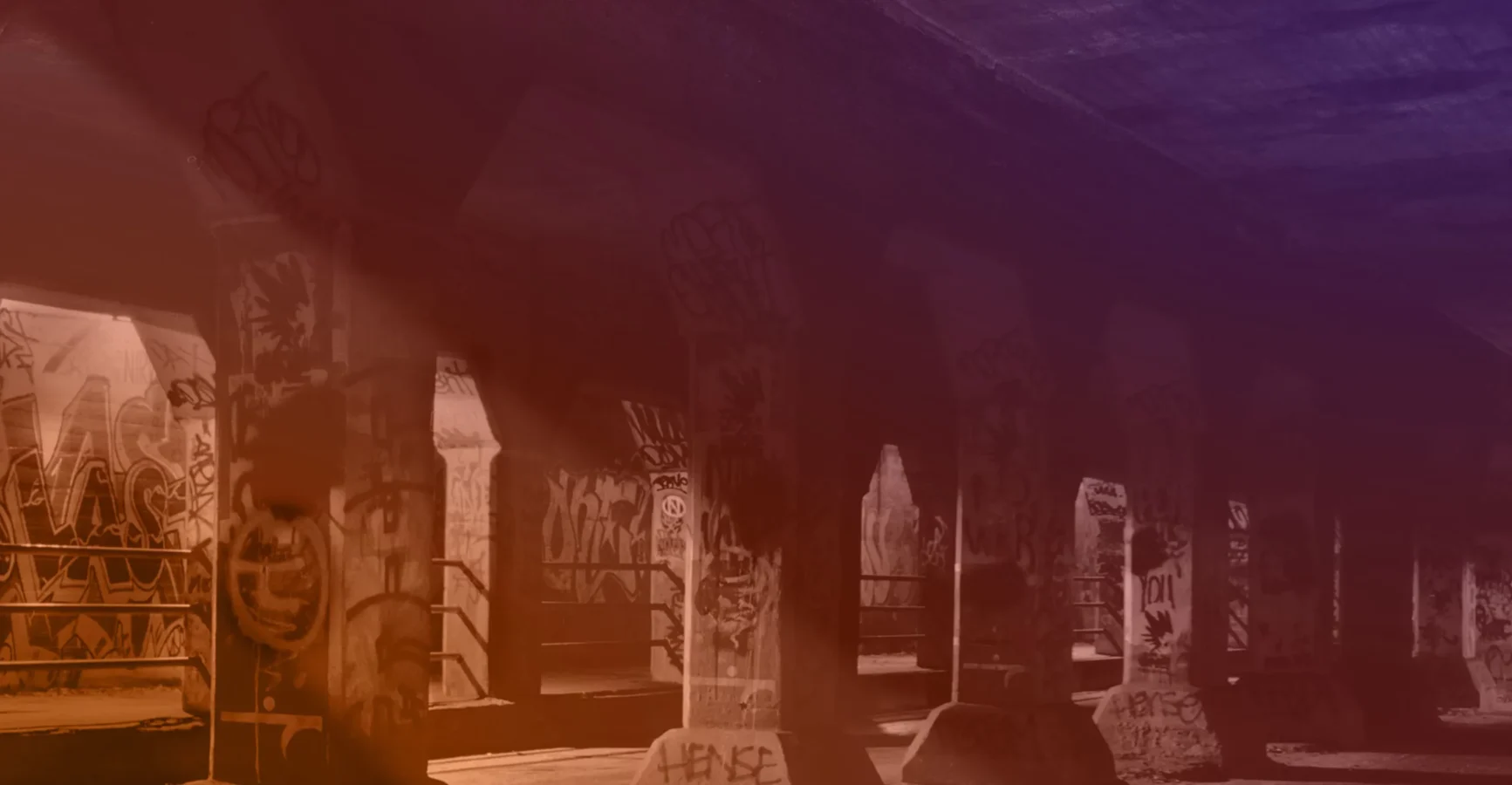 Concrete columns and walls covered in graffiti under a bridge, illuminated by filtered sunlight.