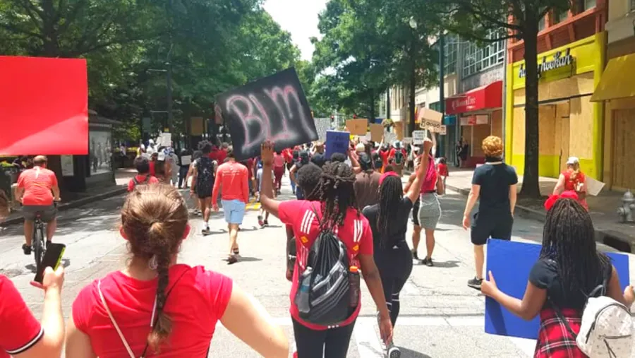 A group of people march down a city street holding signs, including one with "BLM" written on it; many participants wear red shirts.