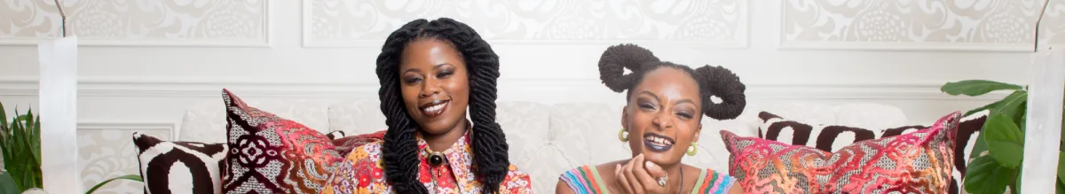 Two women sit on a sofa with colorful patterned pillows, smiling at the camera in a bright room with ornate white walls.
