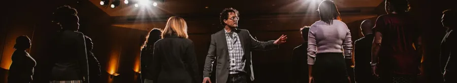 A man in a blazer gestures while standing among a group of people on a dimly lit stage with spotlights overhead.