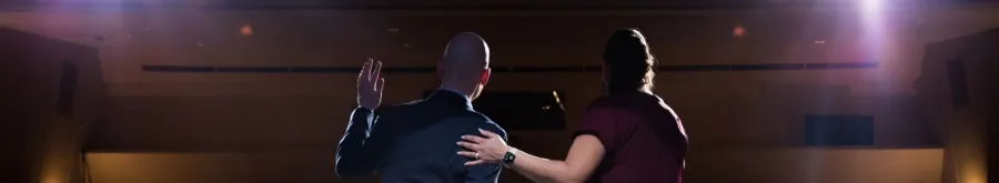 Two people, seen from behind, stand on a stage facing an audience, with bright lights shining towards them.