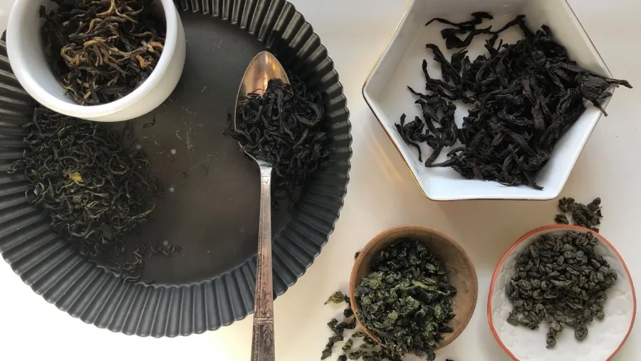 Five different types of loose leaf tea are displayed in various bowls and plates, with a spoon holding some tea leaves on a dark round plate.