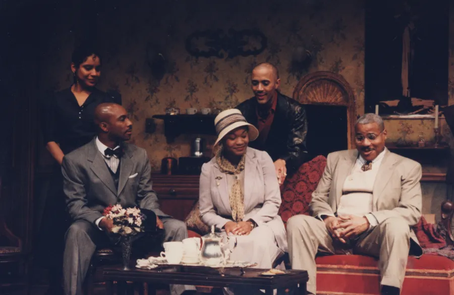 Five people are seated and standing around a tea table in a warmly lit, vintage-style living room set, engaged in conversation.