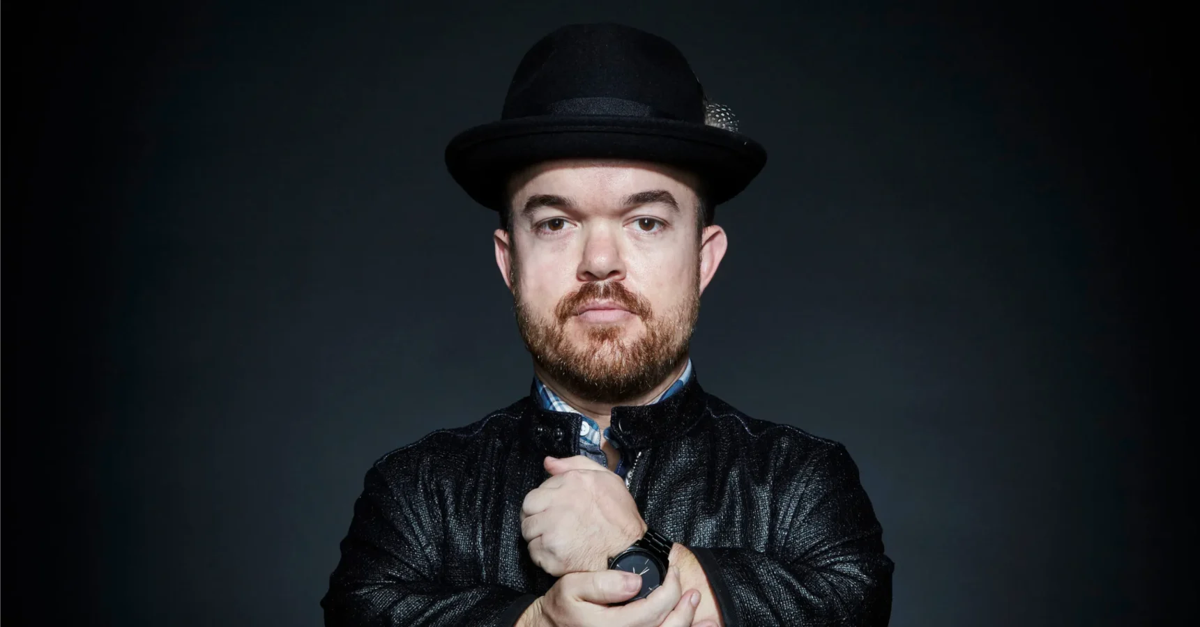 A man with a beard and mustache wearing a black hat and black jacket stands against a dark background, looking directly at the camera with his hands clasped in front of him.