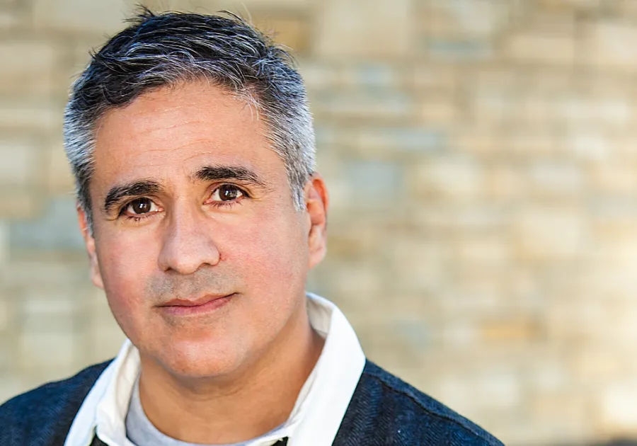 A middle-aged man with short gray hair and a neutral expression is standing in front of a blurred stone wall background.