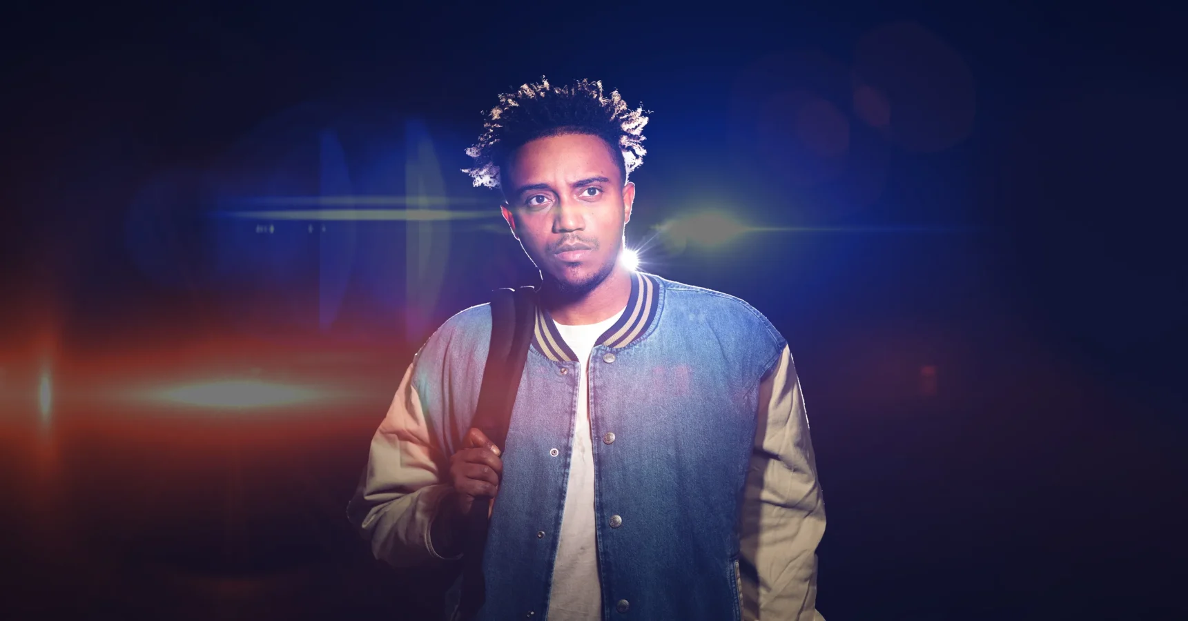 A man wearing a blue and white varsity jacket stands facing forward, holding a bag strap over his shoulder, with bright lights shining behind him.