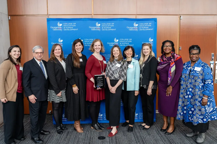 A group of ten people stand smiling in front of a blue College of Education & Human Development backdrop; one woman holds an award plaque.