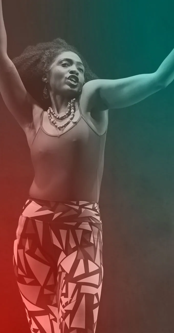 A woman with curly hair raises her arms while performing on stage, wearing a sleeveless top, patterned pants, and layered necklaces.