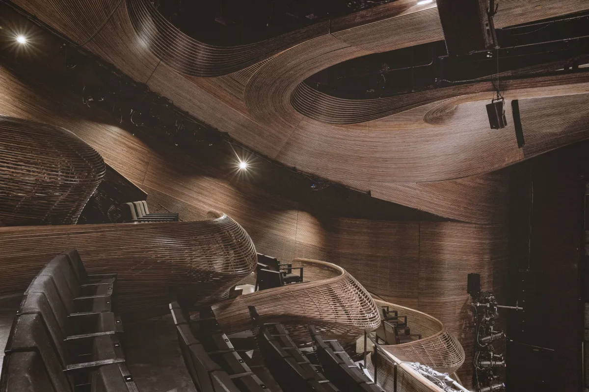 A modern theater interior features sculpted wooden walls and ceiling with flowing curves, black seating, and stage equipment visible to the side.