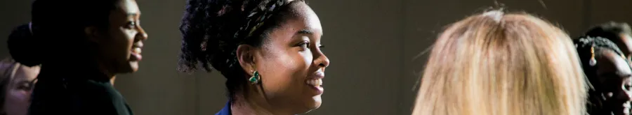 A young woman with braided hair and earrings is smiling and looking to the right, surrounded by other people in a dimly lit indoor setting.